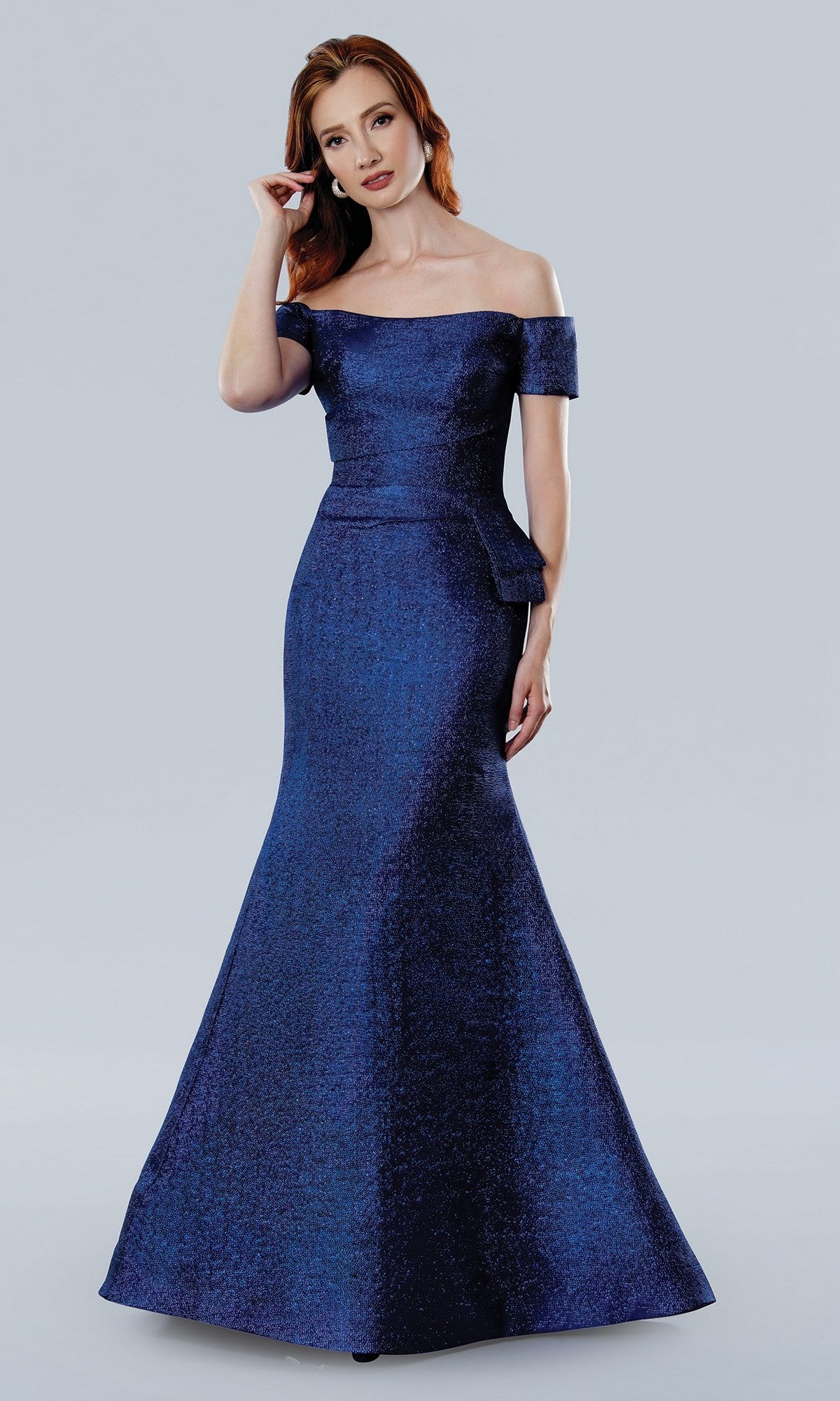 A woman wears the Stella Couture 25331 long formal dress in royal, featuring an off-the-shoulder design and a fitted silhouette that flows to the floor, set against a plain light gray background.
