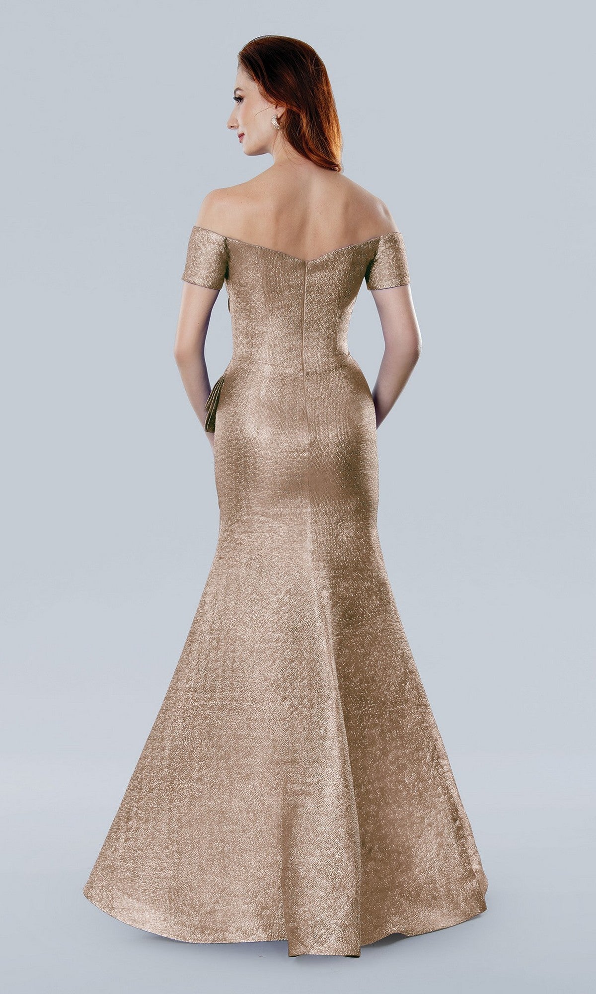 A woman faces away from the camera in a rose Stella Couture 25331 long formal dress, its off-the-shoulder, floor-length fitted silhouette subtly shimmering under the light against a plain backdrop.