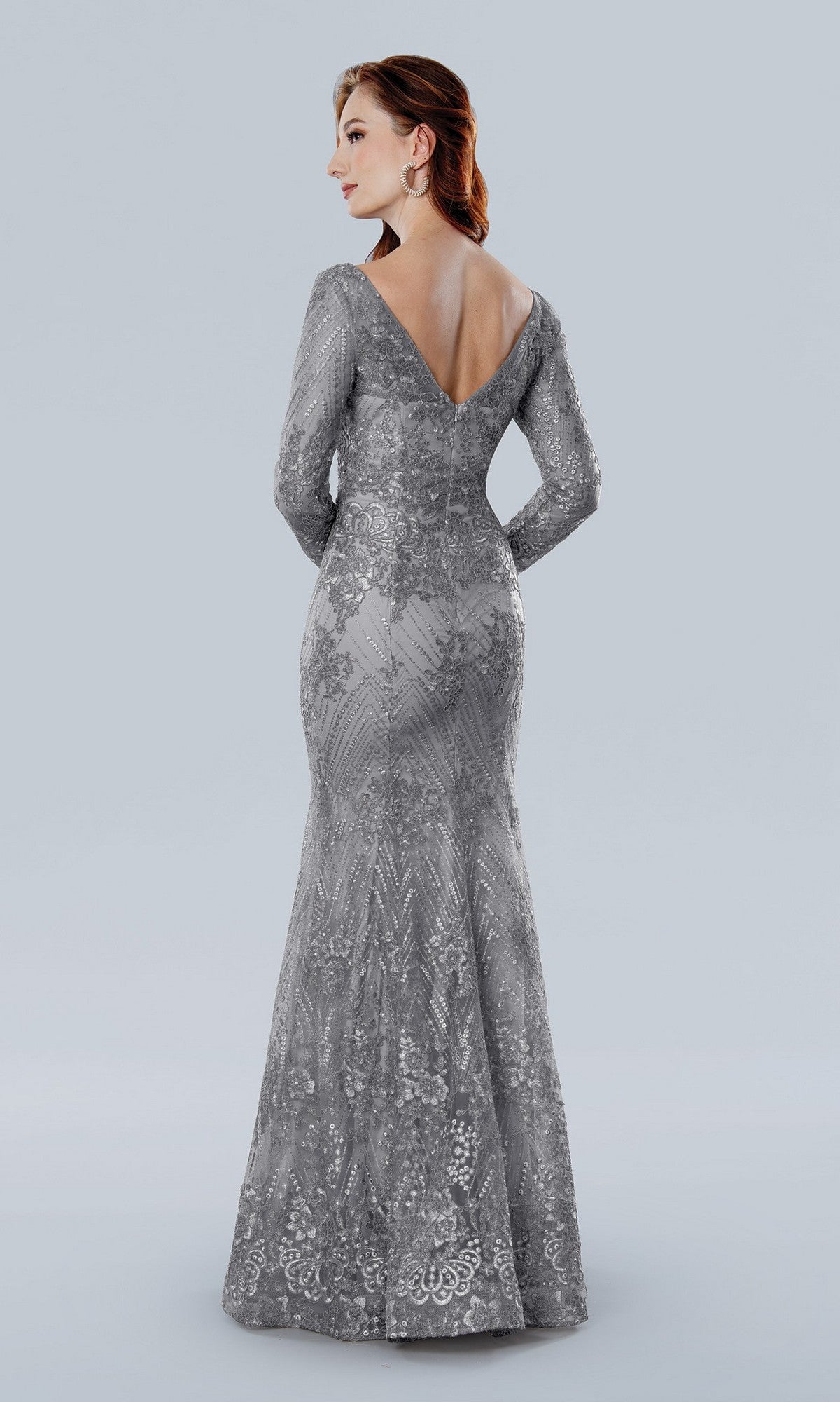 A woman faces away, wearing the Stella Couture 25332 long formal dress in silver—a floor-length gown with sheer sleeves, a V-shaped back, and intricate lace detailing.