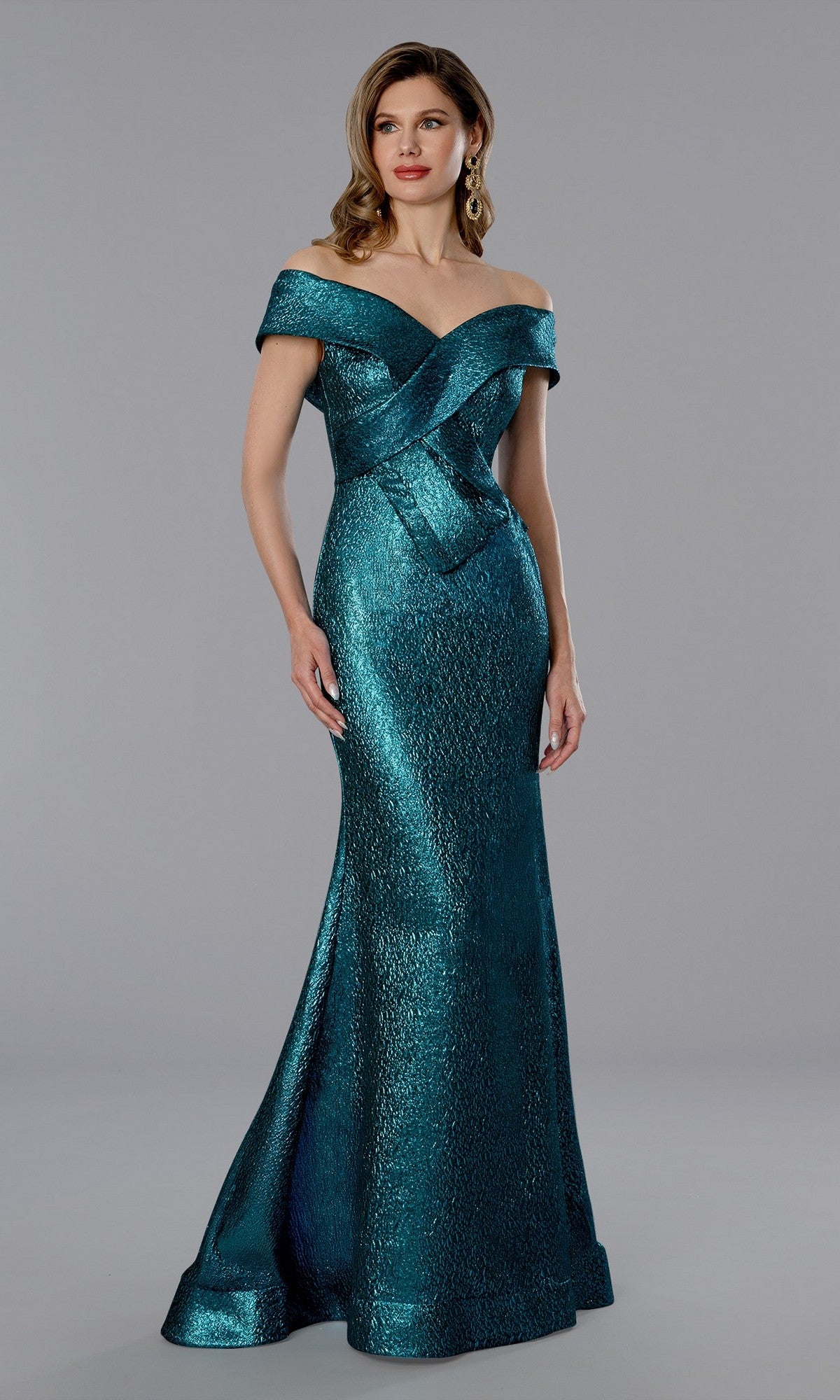A woman poses against a gray background in the Stella Couture 25365 long formal dress—a teal, floor-length, off-the-shoulder mermaid gown with a fitted bodice and flared skirt.