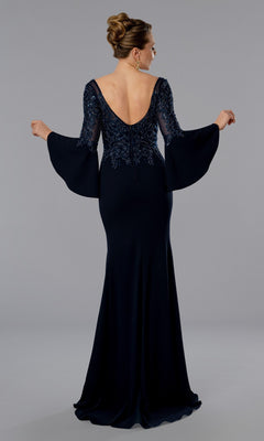 A woman stands against a plain grey background wearing the Stella Couture 25366, a long formal evening gown in navy blue with flared sleeves, elegant lines, and an open back as she faces away from the camera.