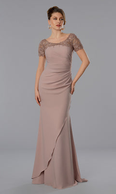 A woman wearing the blush Stella Couture 25368 long formal dress, featuring short beaded lace sleeves, a fitted bodice, and draped details, stands against a simple gray background.