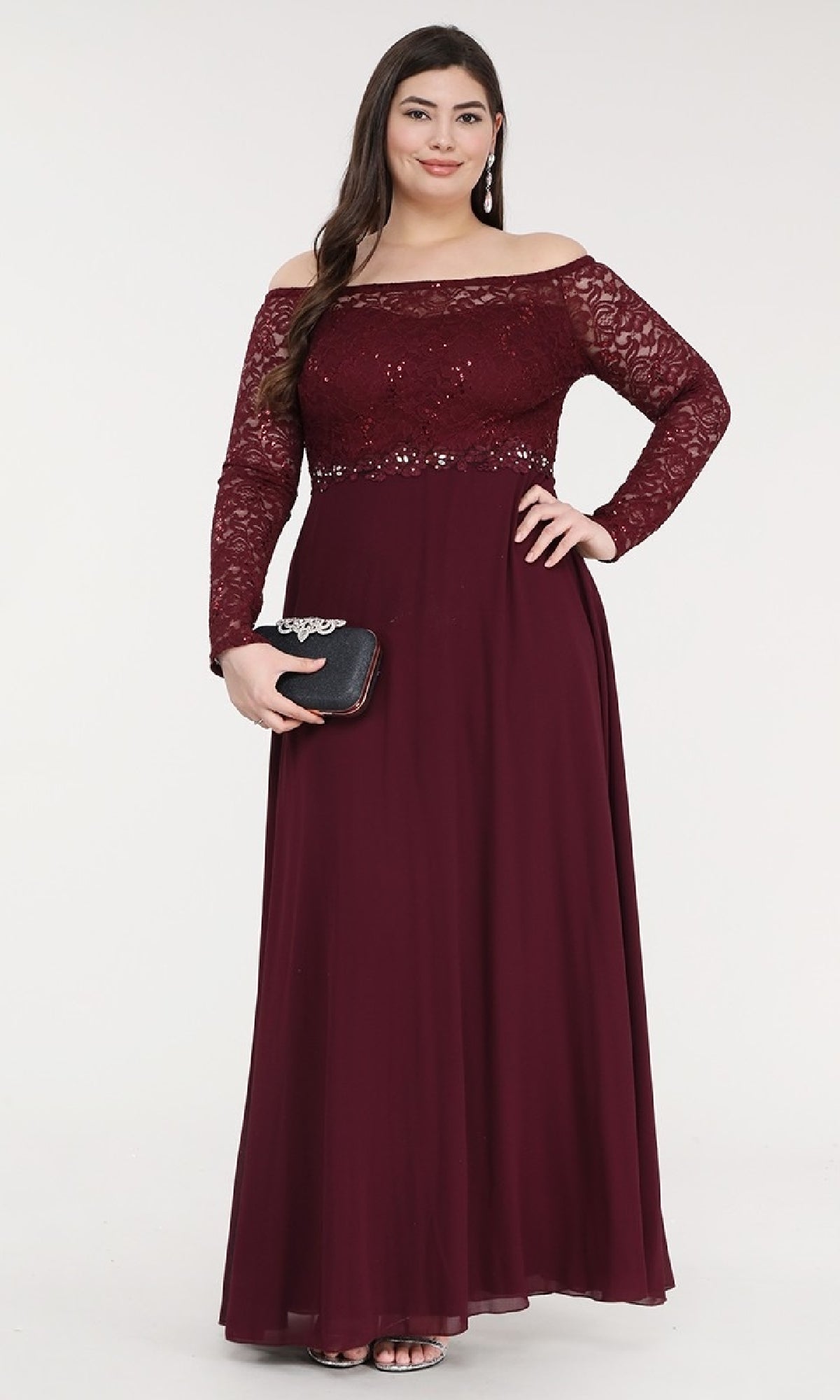 A woman stands against a plain white background holding a black clutch, wearing the Long A-Line Lace-Bodice Plus-Size Formal Dress—an elegant off-shoulder burgundy gown with lace bodice and sleeves and an A-line skirt.