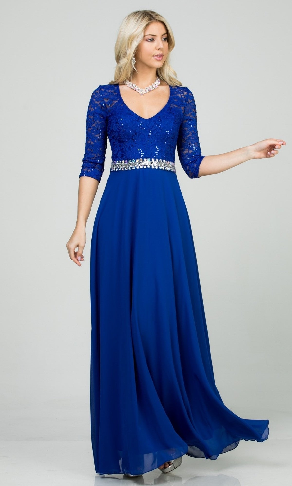 A woman with blonde hair poses gracefully in the Sleeved V-Neck Long A-Line Formal Dress 25434, featuring royal blue sequin lace, half sleeves, a chiffon skirt, and a rhinestone-embellished waistline against a plain background.