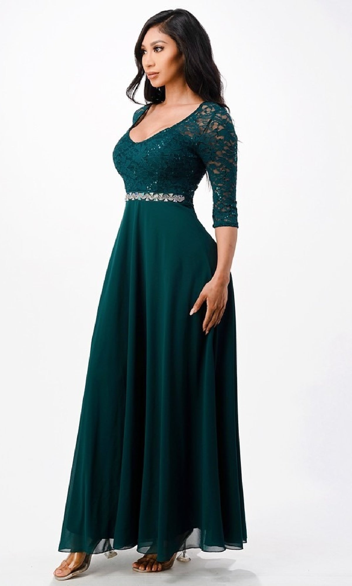 Woman wearing Sleeved V-Neck Long A-Line Formal Dress 25434 in dark green sequin-lace with lace sleeves, a fitted bodice, and rhinestone waist detail, standing against a plain white background.