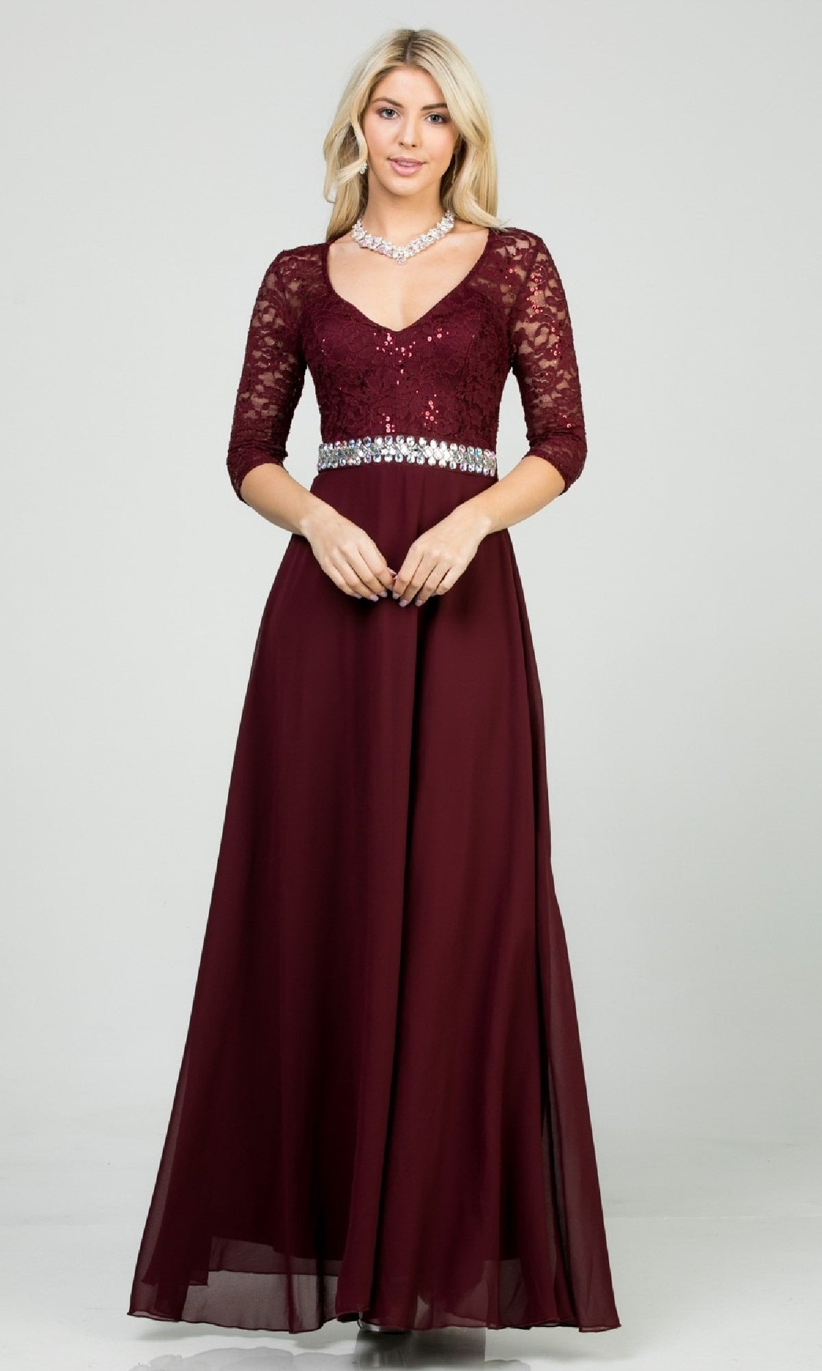 A blonde woman wears the Sleeved V-Neck Long A-Line Formal Dress 25434, featuring a sequin-lace bodice, three-quarter sleeves, rhinestone waistline, and chiffon floor-length skirt. She also accessorizes with a necklace.