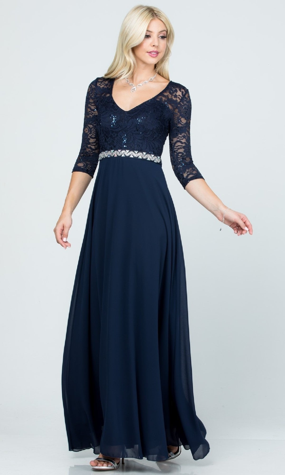 A woman wearing the Sleeved V-Neck Long A-Line Formal Dress 25434, featuring navy blue sequin-lace, three-quarter sleeves, a rhinestone waistline, and a flowing chiffon skirt.
