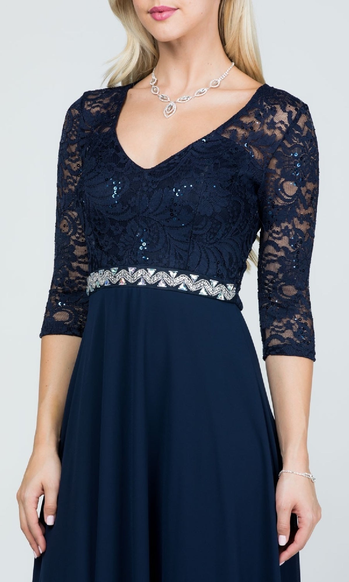 A woman wears the Sleeved V-Neck Long A-Line Formal Dress 25434 in navy blue sequin lace with a chiffon skirt and rhinestone waistband, accessorized with a silver necklace and bracelet.