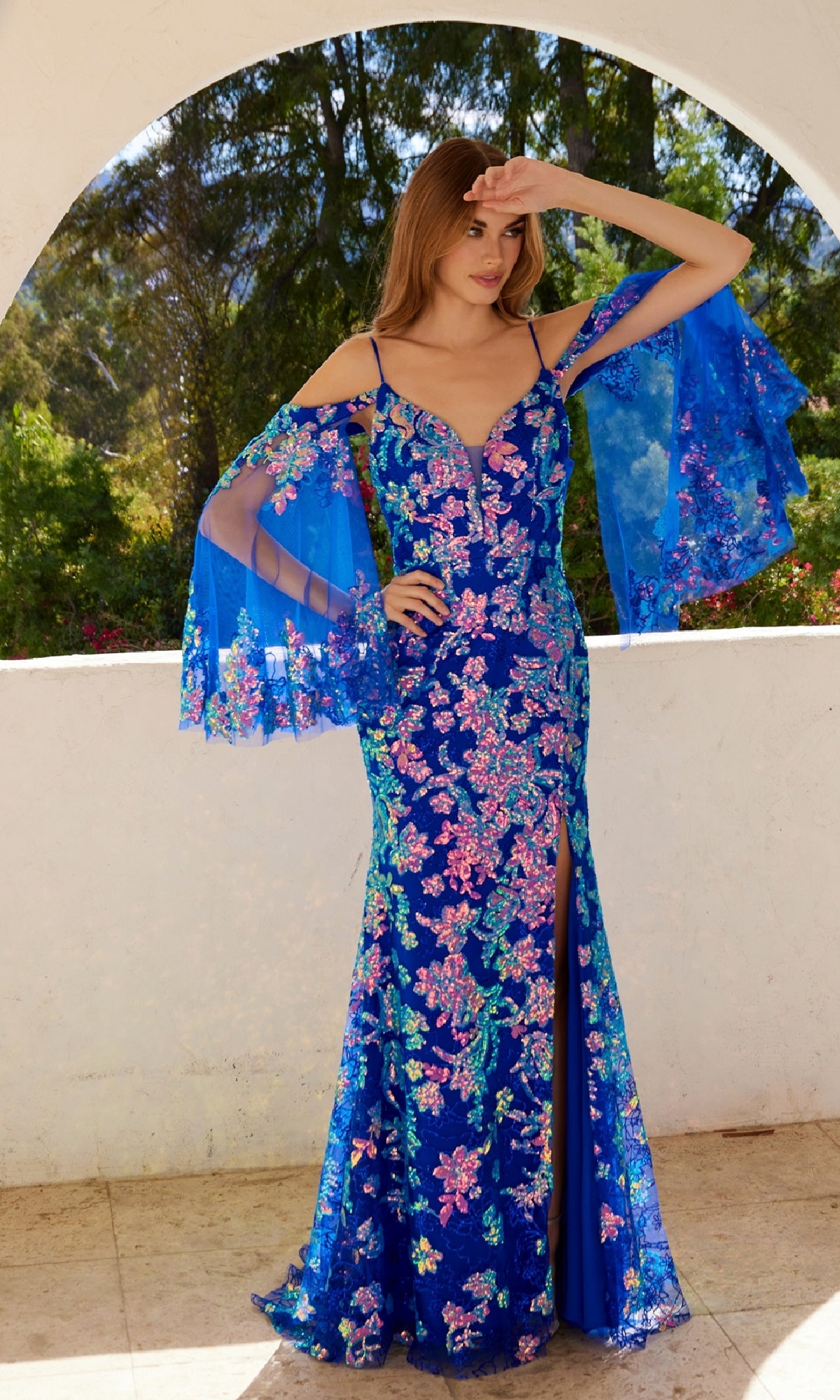 A woman stands on a patio with a garden behind her, wearing the Juliet JT2564U Cape-Sleeve Long Print Prom Dress—a vibrant blue floral gown featuring sheer cape sleeves and a thigh-high slit.