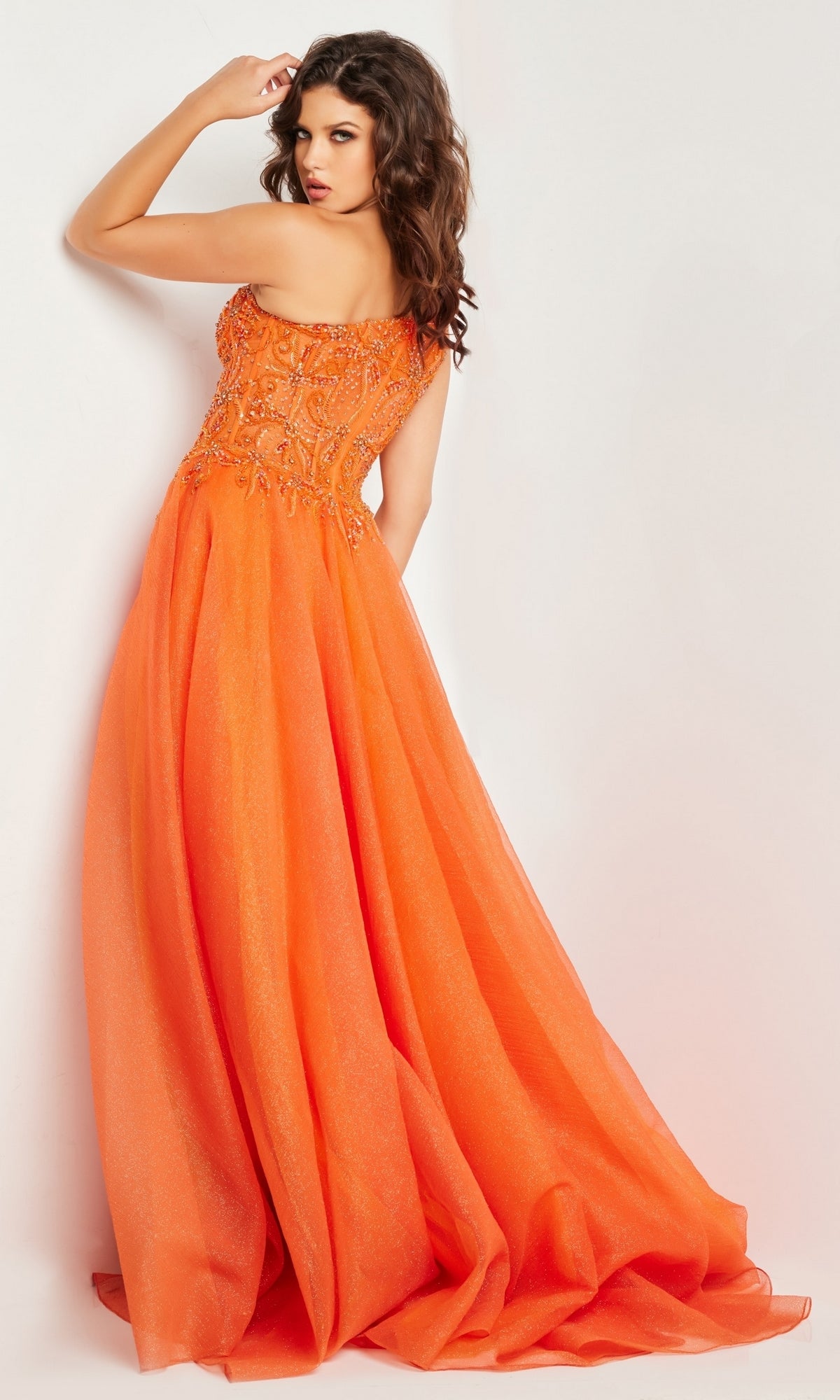A woman in the Jovani One-Shoulder Long Prom Ball Gown 25688, a striking floor-length orange dress with an embellished bodice, stands against a white background—an elegant choice for any special occasion.