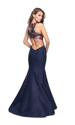 A woman models the La Femme 25885 Navy Blue Long Prom Dress, featuring a sleeveless, backless design with floral embroidery on the bodice and a solid dark blue mermaid skirt, against a white background.