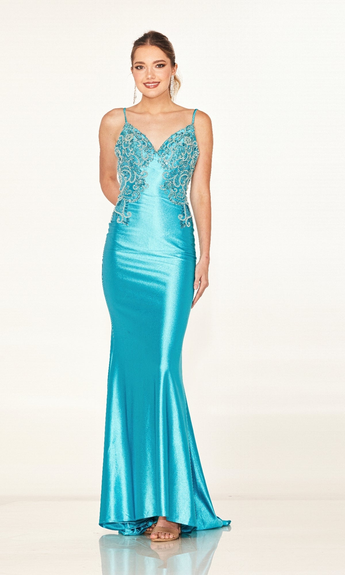 A woman stands against a plain light background, wearing the Cecilia Couture 2602 long prom dress—a form-fitting, shiny turquoise gown with thin straps and ornate bodice detailing.