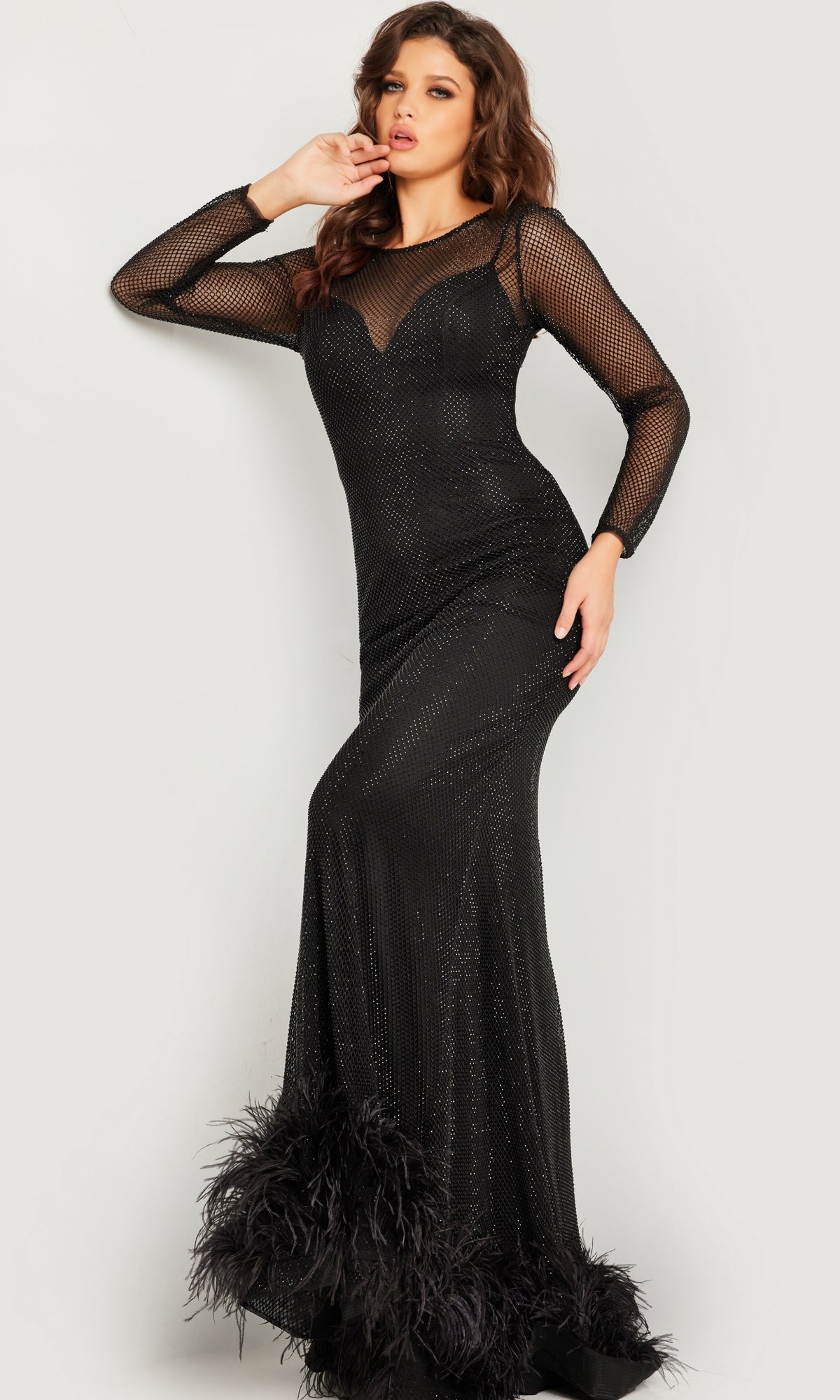 A woman models the Jovani Long Sleeve Long Black Formal Dress 26047, featuring mesh sleeves and feather-accented hem, highlighting the gown's elegant designer style against a plain light background.