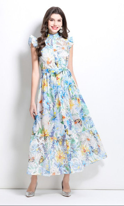 A woman stands against a white background, smiling in a baby blue A-Line Floral-Print Casual Maxi Dress with buttons and ruffled shoulders, styled with silver pointed-toe shoes.