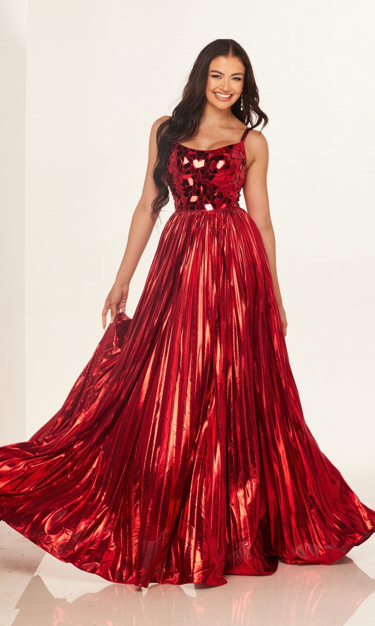 A woman wearing the Cecilia Couture 2609, a shiny red, floor-length pleated A-line prom dress with an embellished bodice and flowing skirt, stands against a plain white background.