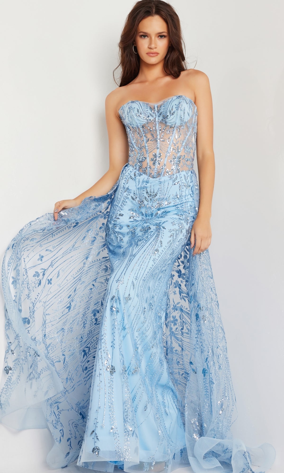 A woman models the Jovani Strapless Long Glitter-Sequin Prom Dress 26113, featuring a sheer corset bodice, floral accents, and a flowing overskirt, standing before a plain light background.