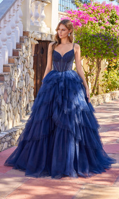 A woman stands outdoors in sunlight wearing the Long Prom Dress: Juliet JT2611N, a dark blue strapless gown with a fitted bodice, surrounded by stone walls and blooming flowers.