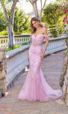 A woman in a baby pink Juliet JT2612N long prom dress with off-the-shoulder mermaid silhouette, sheer corset bodice, and floral appliqué stands on a stone walkway in a garden.