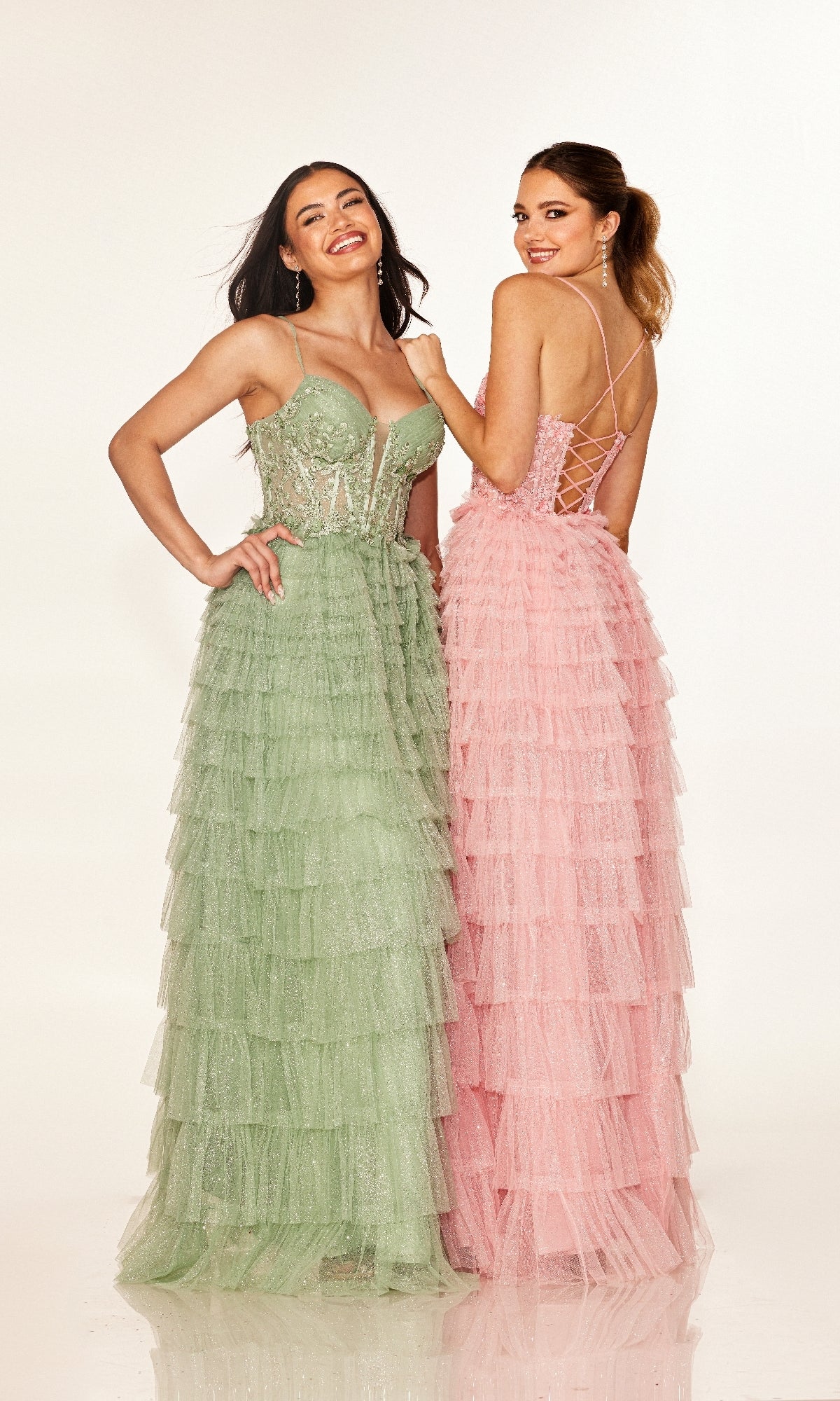 Two women stand side by side, smiling, in floor-length tiered dresses—one wears the Cecilia Couture 2613 long light green prom dress with a fitted bodice, and the other is in pink with a lace-up open back.