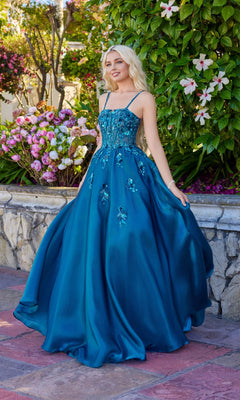 A woman in the teal Juliet JT2617U long prom dress, featuring a lace-up back and embellished A-line design, stands outdoors on a stone patio amid flowers and greenery.