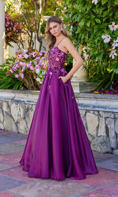 A woman stands outdoors among greenery and pink flowers, wearing the Juliet JT2617U long magenta sleeveless A-line prom dress with floral embellishments.