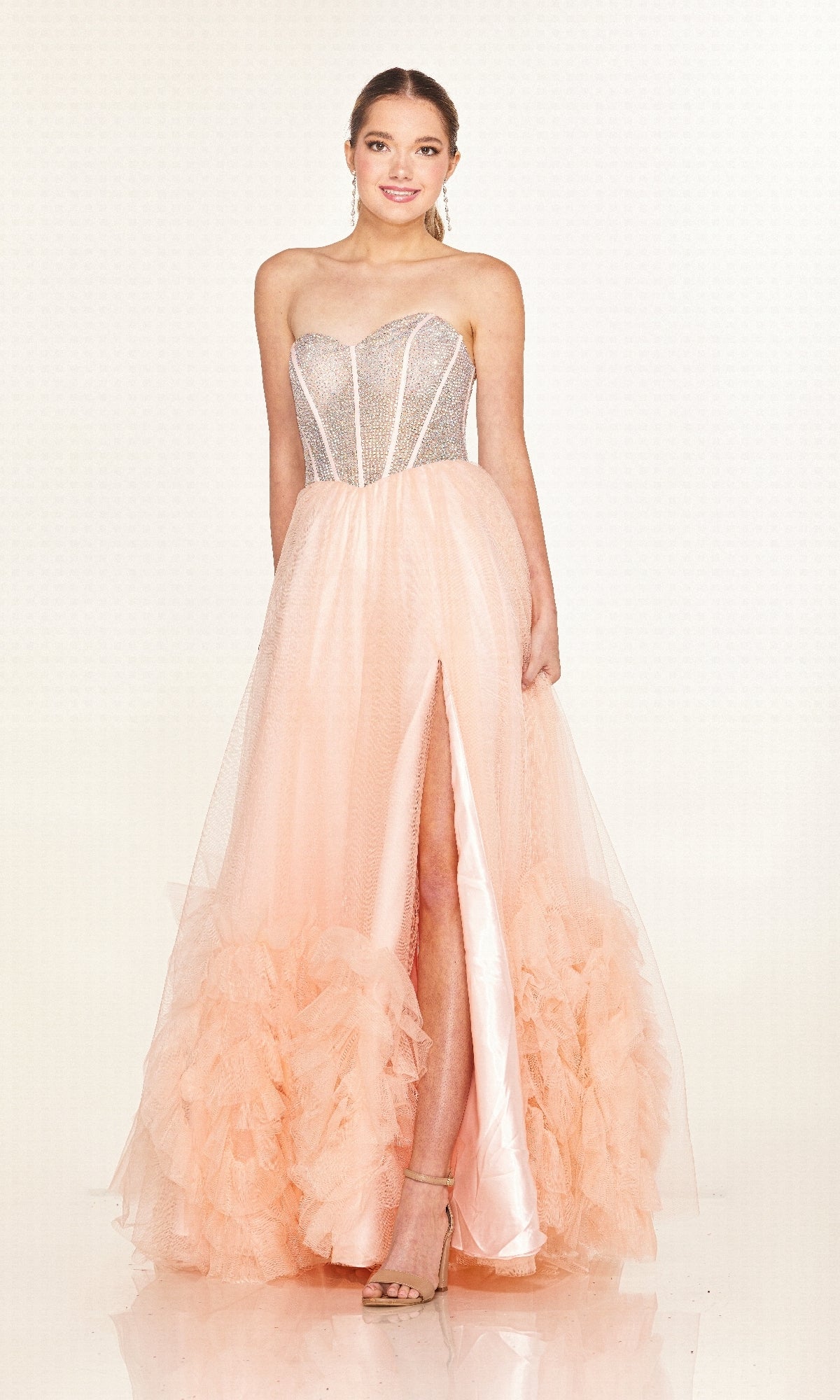 A woman models the Cecilia Couture 2617, a long strapless prom dress with a rhinestone corset bodice, sheer panels, peach tulle A-line skirt with thigh-high slit, and ruffled hem, set against a plain background.