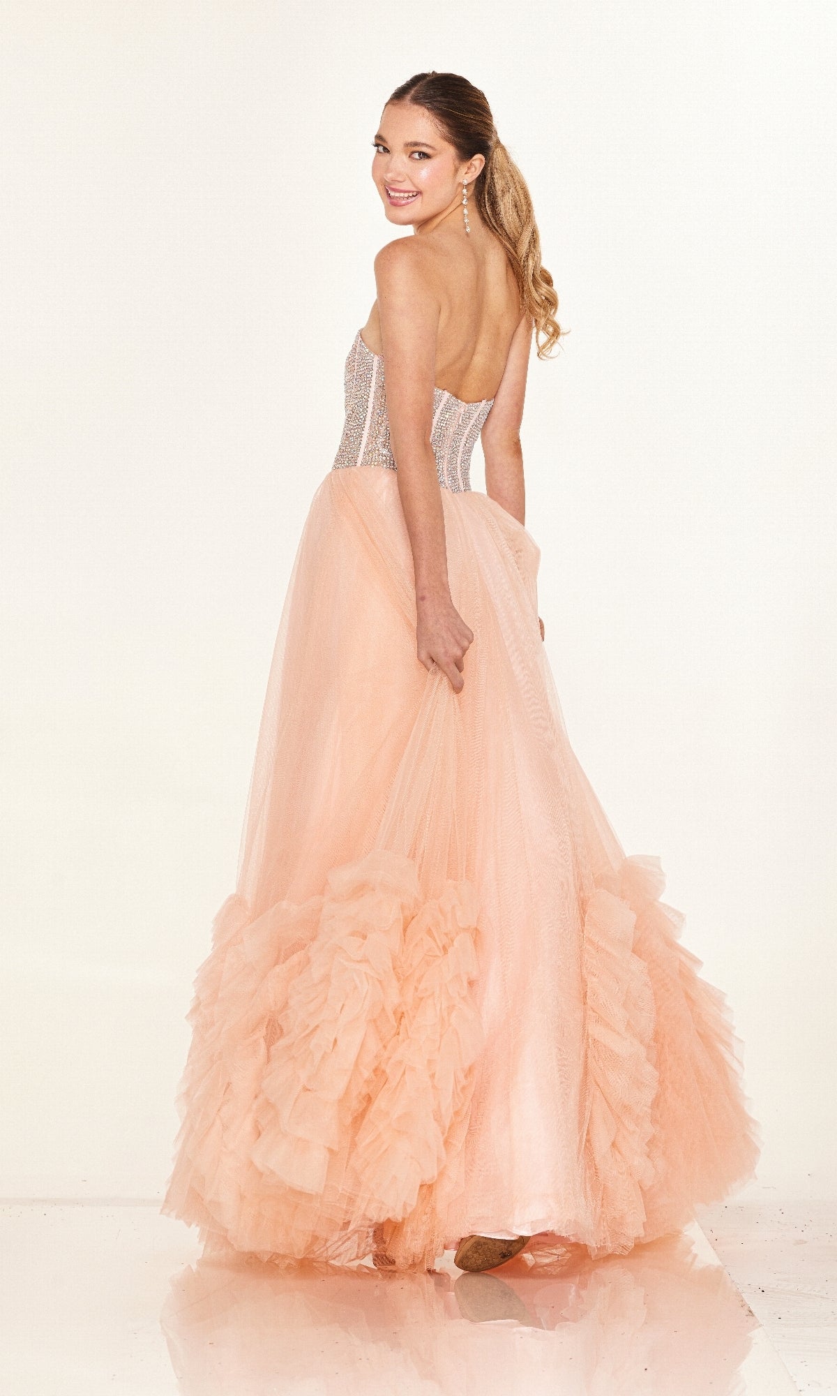 Woman in the Cecilia Couture 2617 long prom dress, featuring a strapless A-line tulle skirt and fitted semi-sheer bodice, smiling over her shoulder against a light background.