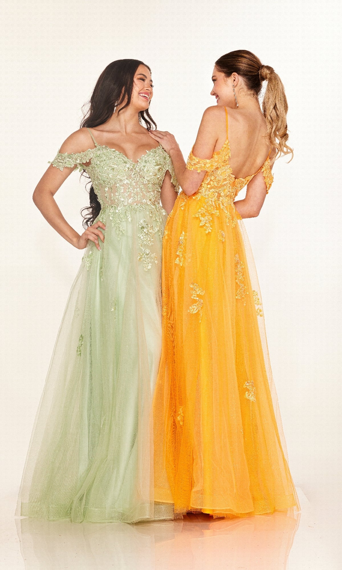 Two women smile at each other—one wears a light green off-the-shoulder prom dress with embellishments, the other a yellow embroidered prom dress with spaghetti straps and open back. Both showcase the Long Prom Dress: Cecilia Couture 2618.