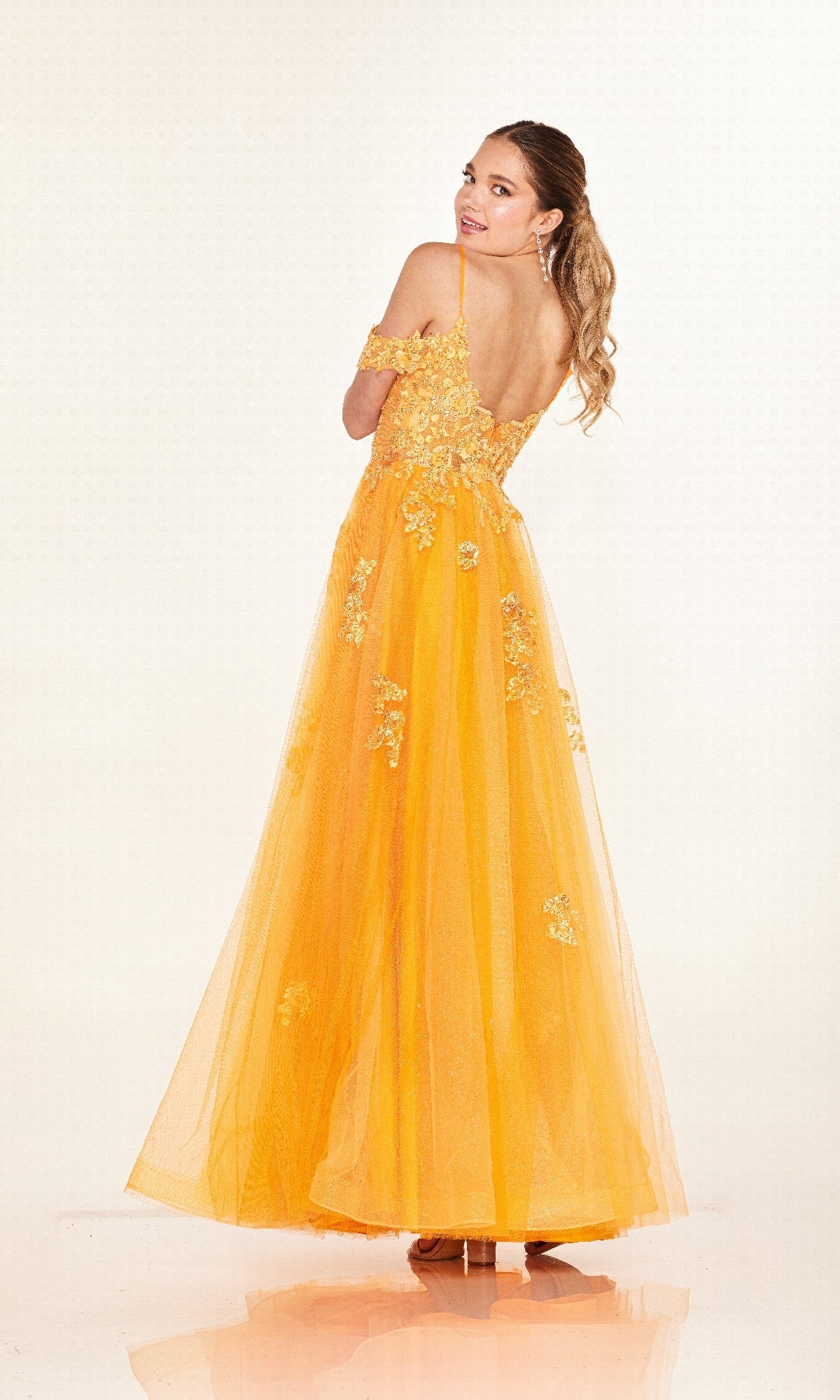 A woman faces backward in the Long Prom Dress: Cecilia Couture 2618, a floor-length bright yellow gown with thin straps, floral embroidery, and embellishments, set against a plain white background.
