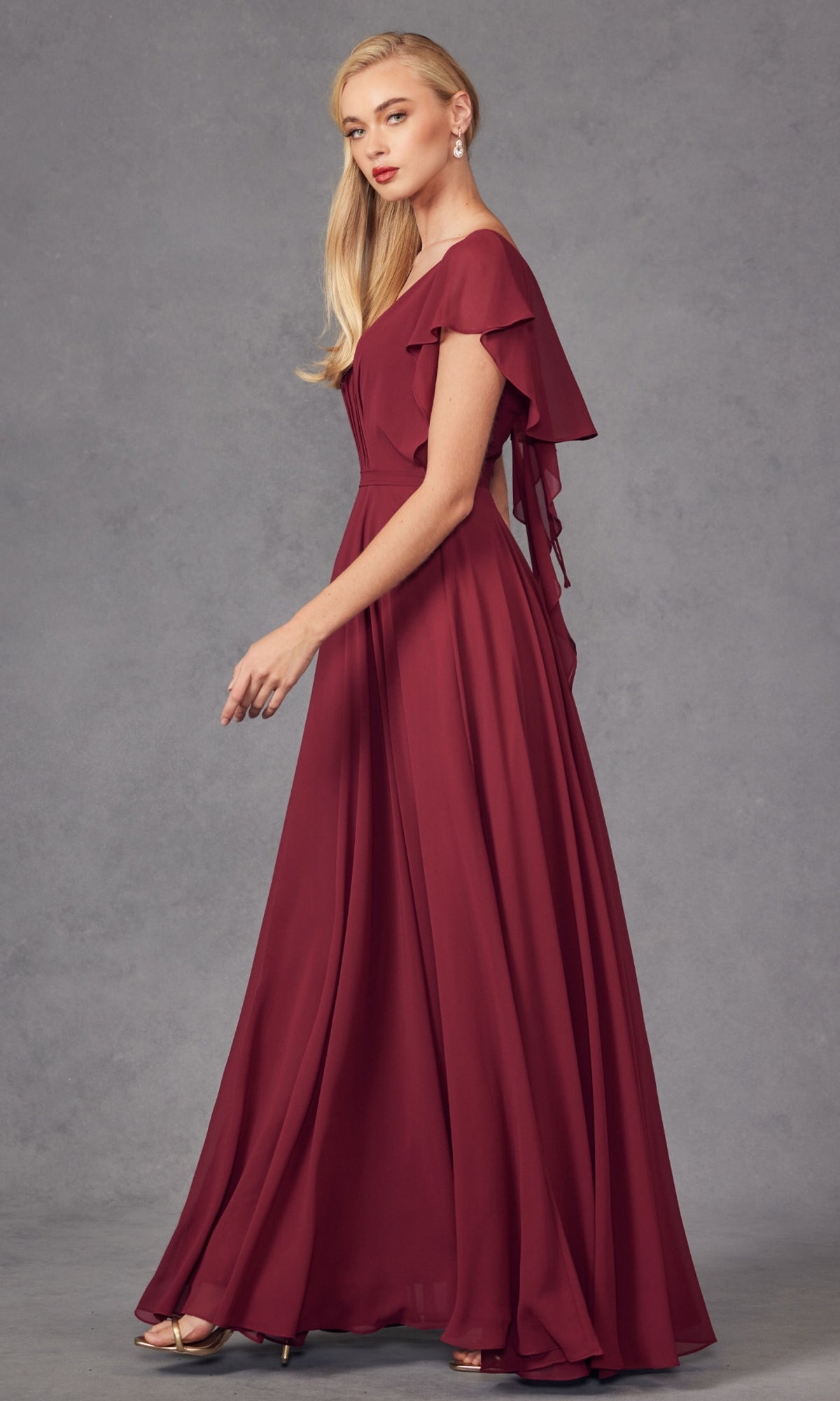 A woman with long blonde hair models the Flutter-Sleeve Long Formal Evening Gown 261, a floor-length burgundy A-line dress with short flutter sleeves and an open back, standing before a plain gray background.