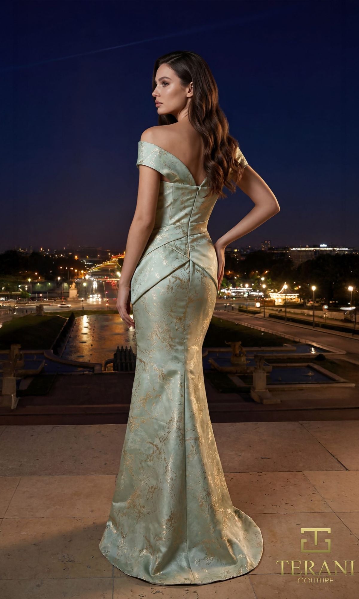 A woman in a light blue Terani 261E6252 off-the-shoulder mermaid gown stands on a terrace at night, city lights twinkling in the background.