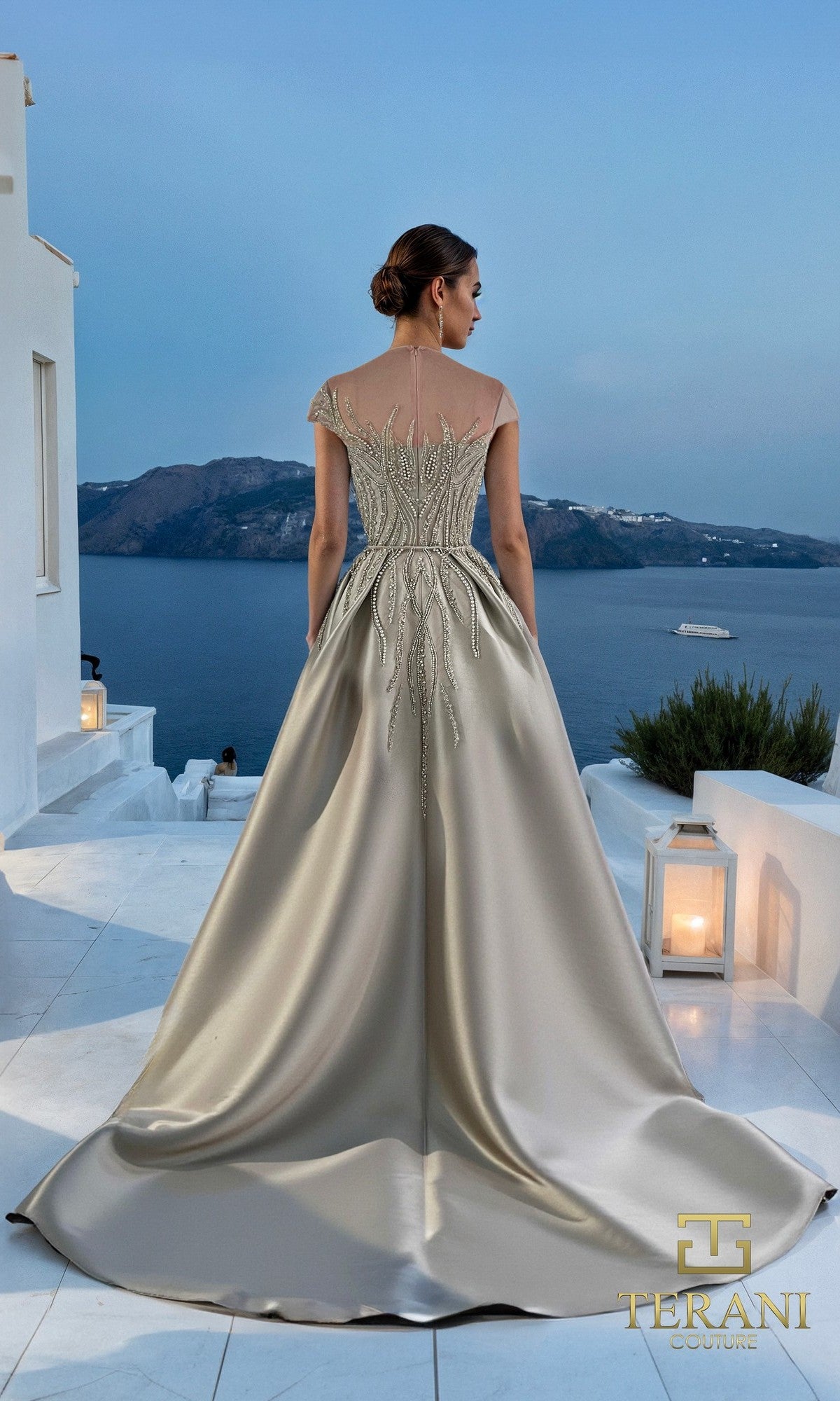A woman wears the Long Formal Dress: Terani 261E6254, made of stretch Mikado with silver embroidery, as she stands on a white terrace overlooking the sea and mountains at sunset.