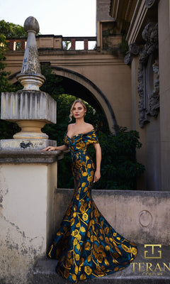 A woman poses by a stone balustrade in front of an ornate historic building, wearing the Long Formal Dress: Terani 261E6273, an off-shoulder, floor-length blue and gold jacquard gown. The "Terani Couture" logo appears in the corner.