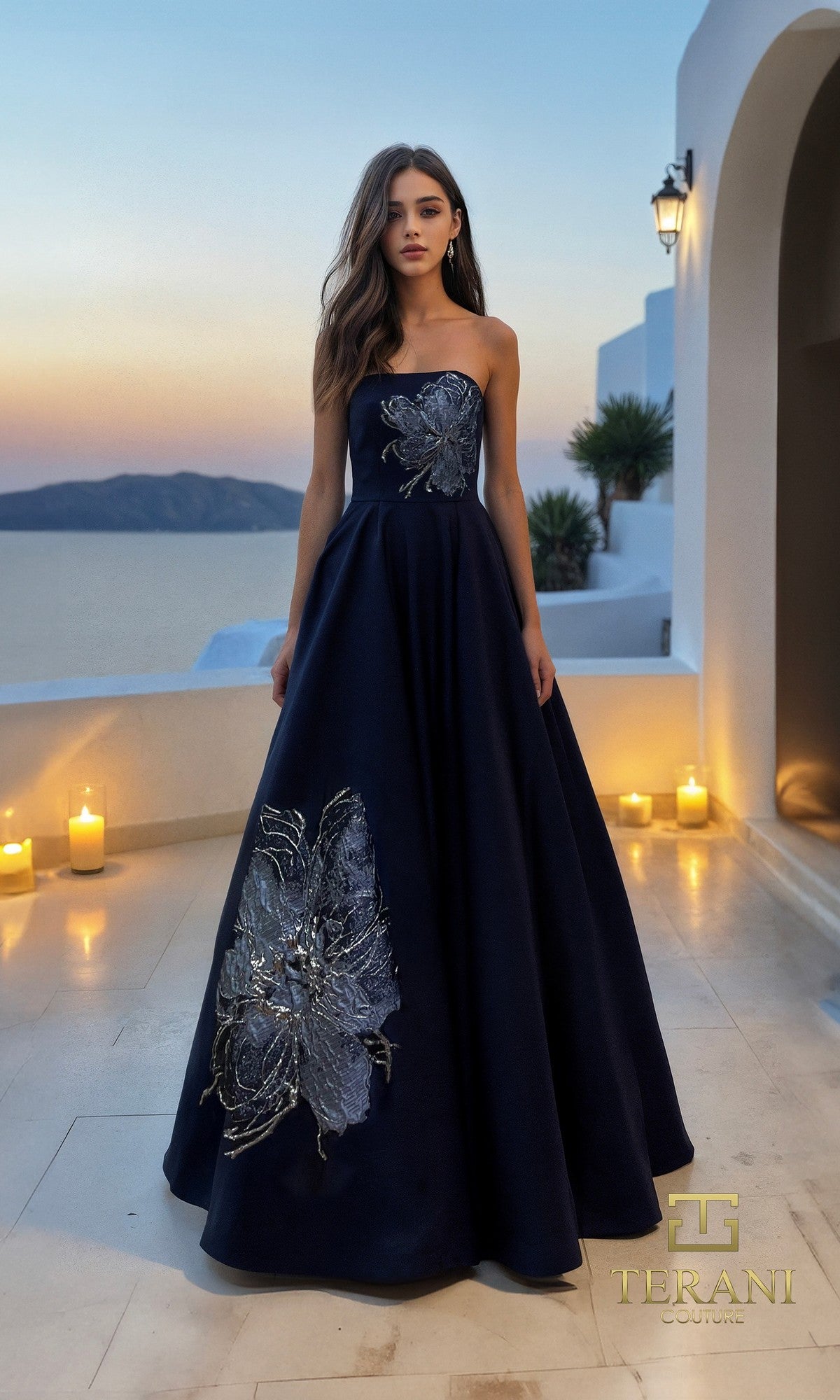 A woman in the navy Long Formal Dress Terani 261E6544, featuring a strapless a-line silhouette and silver floral embellishments, stands on a terrace at sunset surrounded by glowing candles.