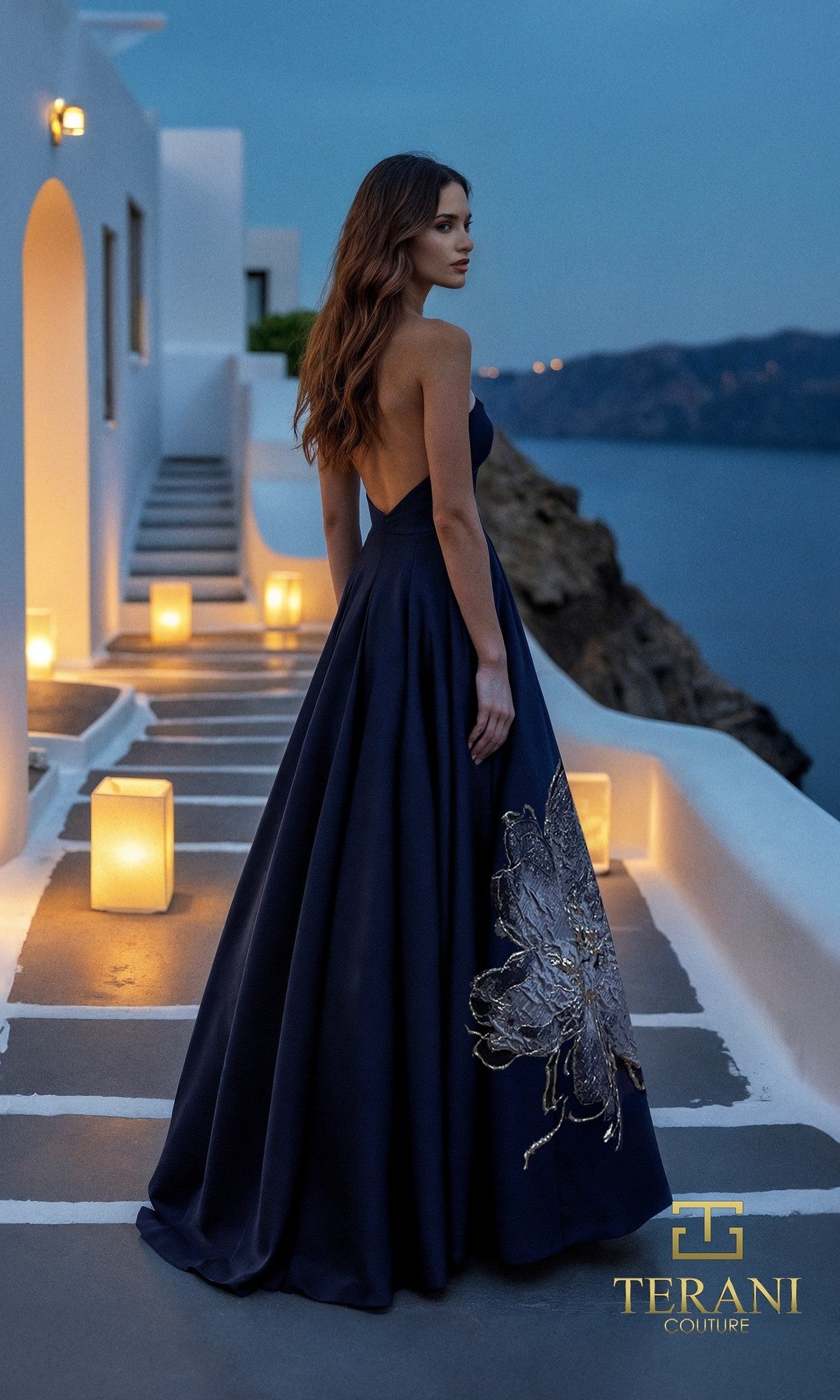 A woman in the Long Formal Dress: Terani 261E6544, featuring a large butterfly design, stands on a coastal terrace at dusk, surrounded by glowing lanterns.