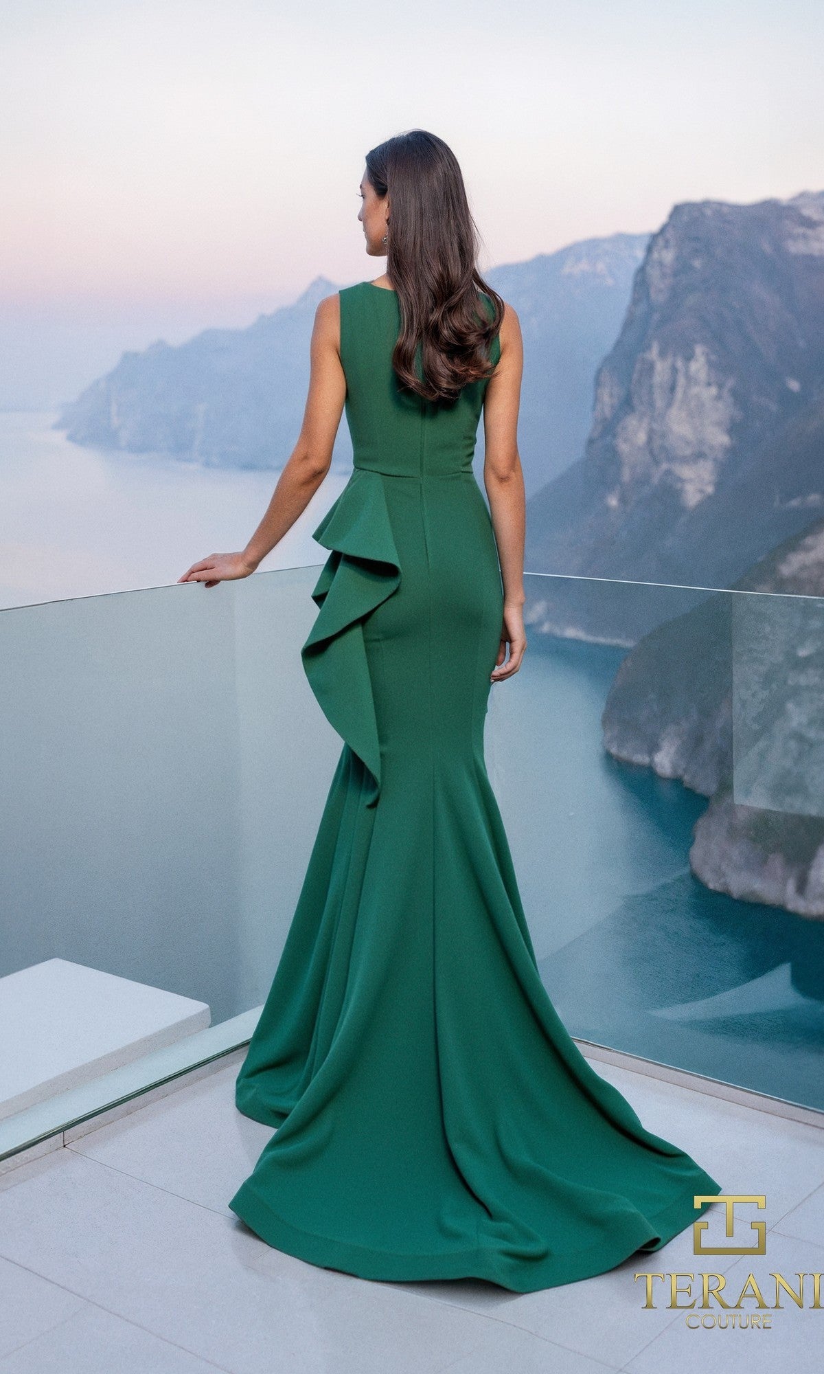A woman in a long Terani 261E6750 formal dress with ruffle detail stands on a balcony overlooking cliffs and water.
