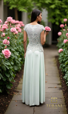 A woman in the Long Formal Dress: Terani 261M6437, featuring a light blue embroidered bodice, stands on a stone path among rose bushes, holding a pink rose and facing away from the camera.