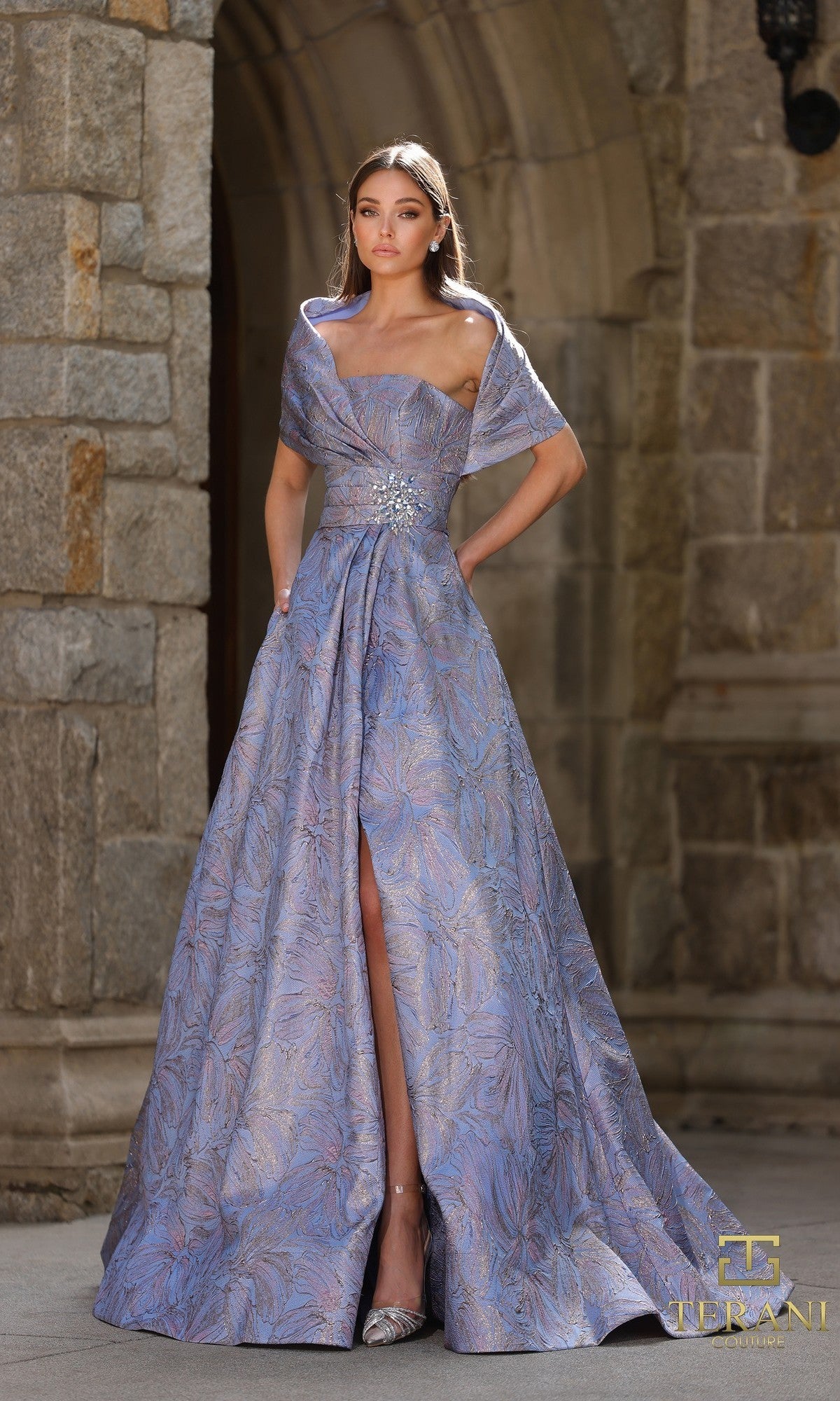 A woman in a Blue Rose long formal dress (Terani 261M6453) with a jeweled brooch and front slit stands gracefully before a stone archway.
