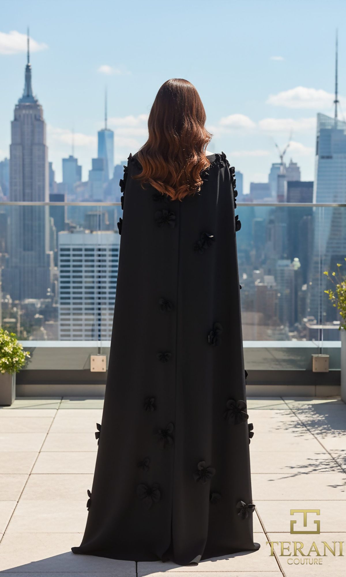 A woman with wavy brown hair stands on a rooftop, facing away, wearing the Long Formal Dress: Terani 261M6687. The city skyline, with the Empire State Building in view, stretches out behind her.