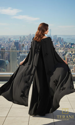 A woman in the Terani 261M6687 long formal dress, featuring a flowing black cape with floral details and a sleek column silhouette, stands on a rooftop terrace overlooking the city skyline.