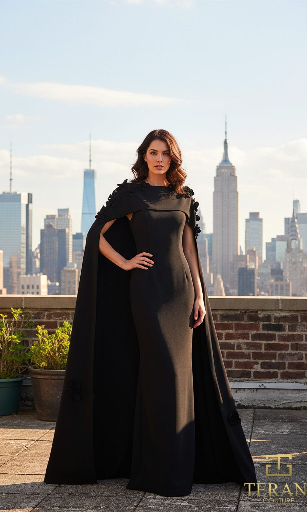 A woman stands on a rooftop in the Terani 261M6687 long formal dress, with its elegant column silhouette and cape, the Empire State Building and city skyline rising behind her?an unforgettable vision of Terani Couture style.