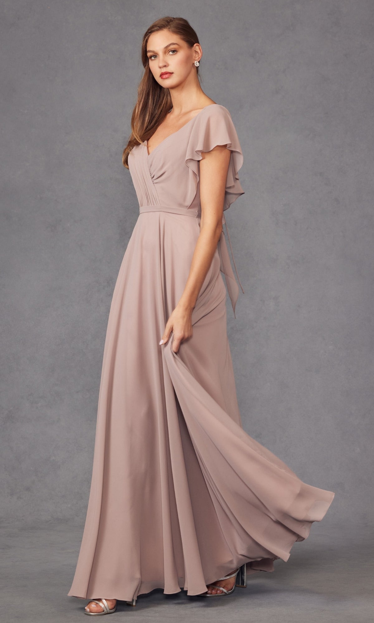 A woman in the Flutter-Sleeve Long Formal Evening Gown 261, a mauve floor-length a-line dress with flutter sleeves, stands against a gray backdrop holding her dress and wearing silver high heels.