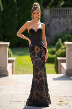 A woman stands outdoors on a stone pathway, wearing the Terani Couture 261P6096 long formal dress in Black Nude. Greenery and a stone wall are in the background, with the Terani Couture logo at the lower right corner.