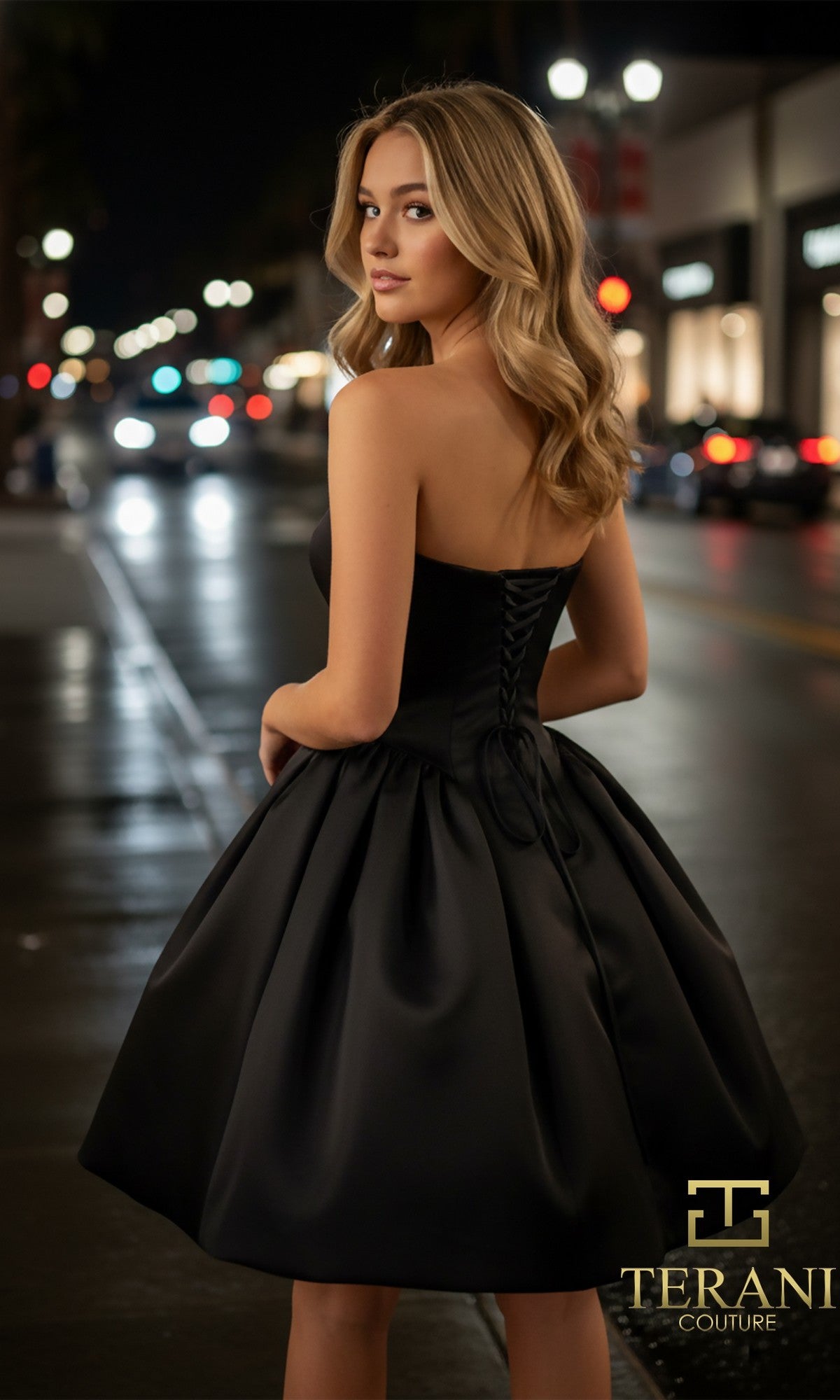 A woman in the Terani 261P6176 short prom dress stands on a city street at night, looking over her shoulder as shop lights and blurred car headlights illuminate the background.