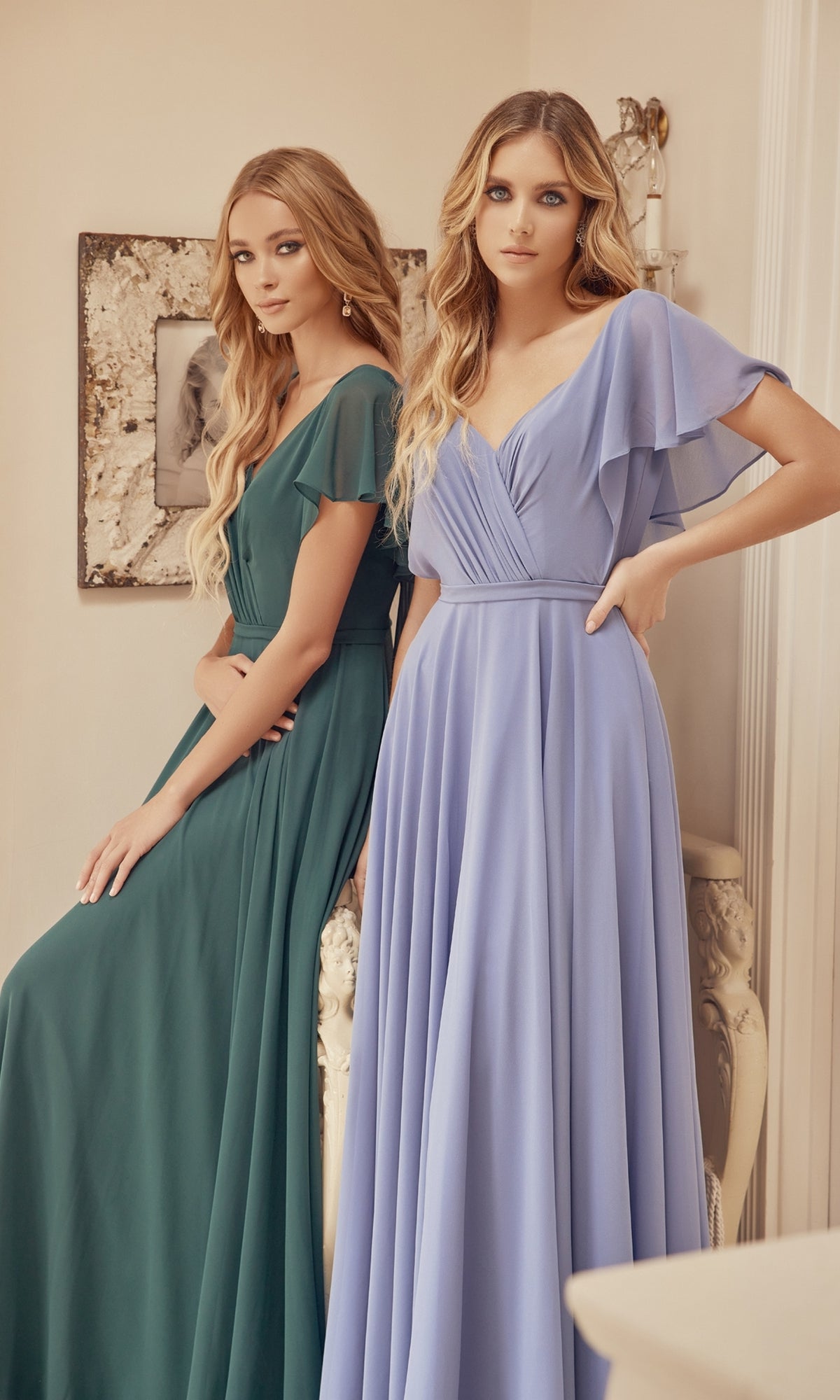 Two women in floor-length gowns—one in the Flutter-Sleeve Long Formal Evening Gown 261, the other in a light blue a-line dress—stand indoors by a decorated white console table and a framed photo on the wall.