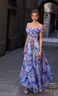 A woman wearing the Long Formal Dress: Terani 261C6641 stands on a narrow stone street, looking at the camera in silver shoes, with the "TERANI Couture" logo visible.