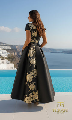 A woman in the Terani Couture Long Formal Dress 261E5194, a black and gold floral gown with a tea-length overskirt, stands by a pool overlooking coastal cliffs and white buildings. The Terani Couture logo is displayed.