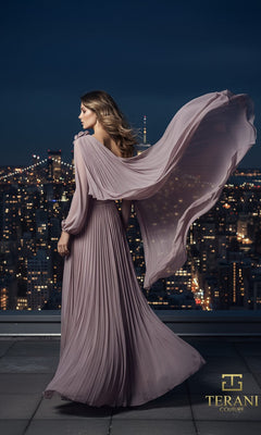A woman in a pleated, lavender one-shoulder chiffon dress (Long Formal Dress: Terani 261E6206) stands on a rooftop at night with city lights behind her, the gown?s cape flowing in the wind. The Terani Couture logo is visible.