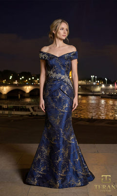 A woman stands outdoors at night in the Terani 261E6252 long formal dress, a floor-length midnight gold gown. City lights and a bridge over water provide a stunning backdrop.