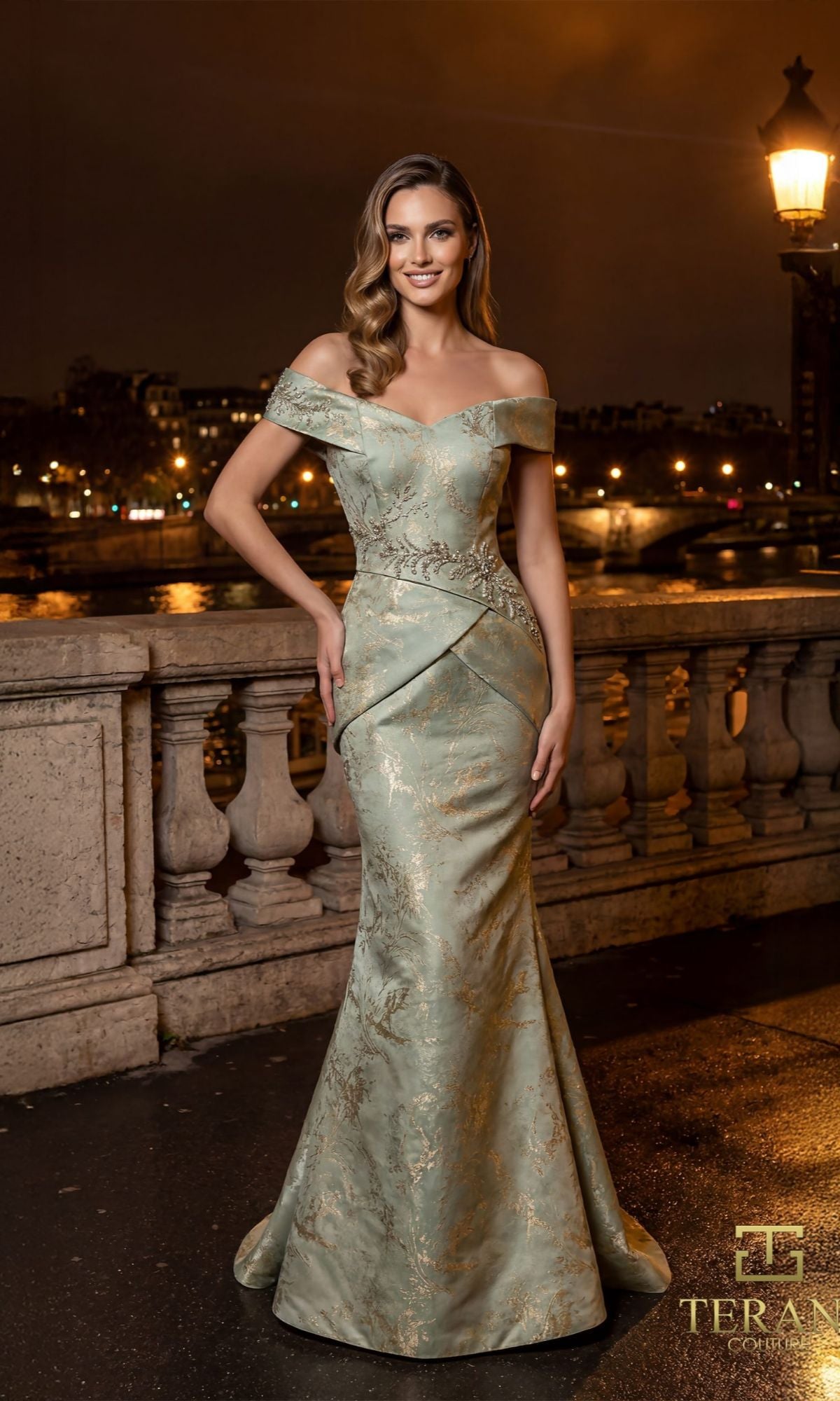A woman in the Terani 261E6252 long formal dress, featuring luxurious jacquard and a mermaid silhouette, stands by a stone railing at night, illuminated by city lights and a street lamp highlighting the gown's gold patterns.
